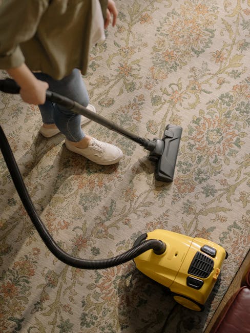 A professional cleaning technician from Barnescray Carpet Cleaners is performing deep surface cleaning on a plush beige carpet in a well-lit, modern living room. The technician is dressed in full prot